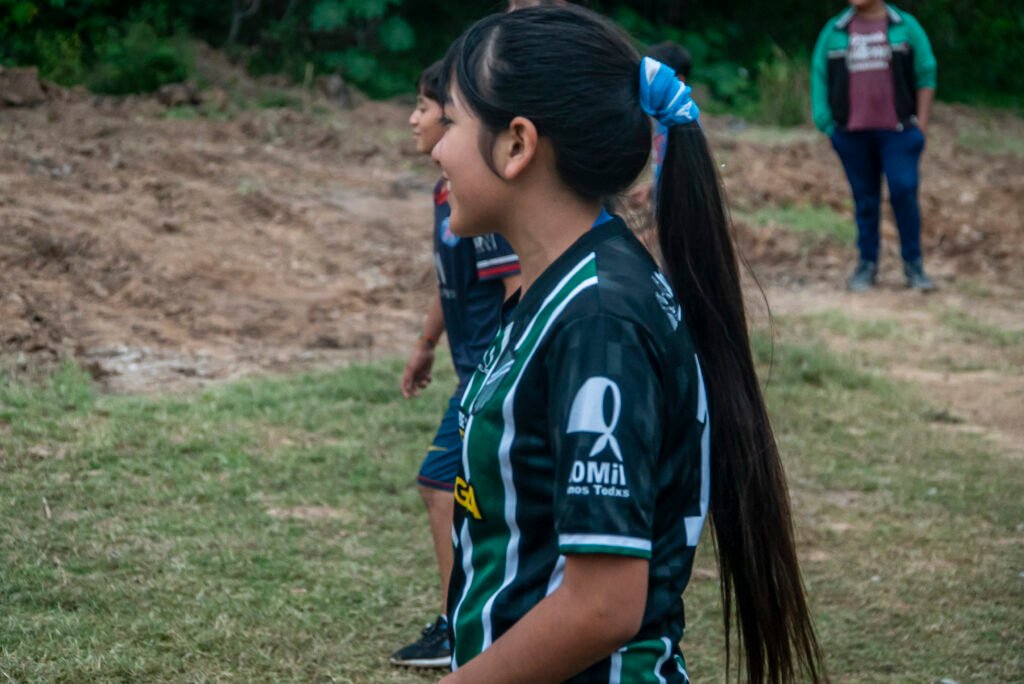 Este imagen muestra a una joven con cabello largo oscuro recogido en una cola de caballo con un coletero azul, vistiendo una camiseta de fútbol negra y verde a rayas.