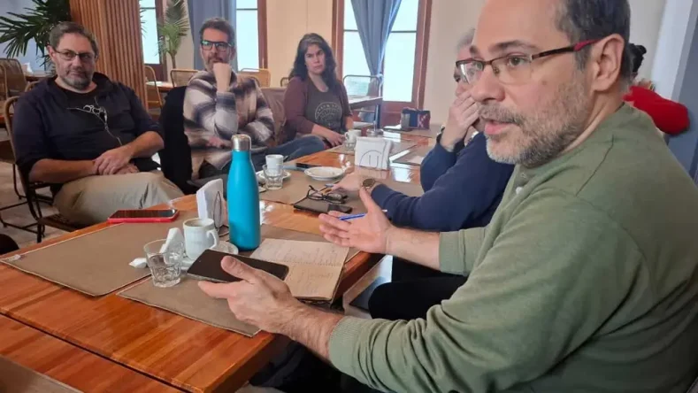 Esta imagen muestra una reunión de trabajo o intercambio entre varias personas sentadas alrededor de una mesa de madera en un espacio interior.