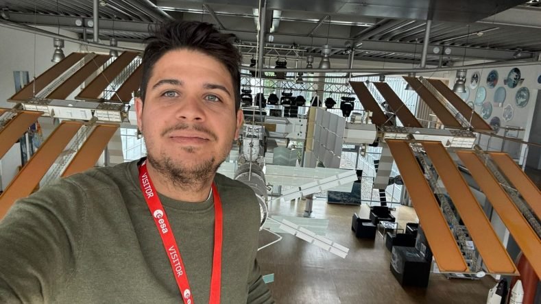 La imagen muestra a un visitante con una credencial de la Agencia Espacial Europea (ESA) tomándose una selfie junto a una maqueta de la Estación Espacial Internacional (ISS) en un centro de exhibición de la ESA.