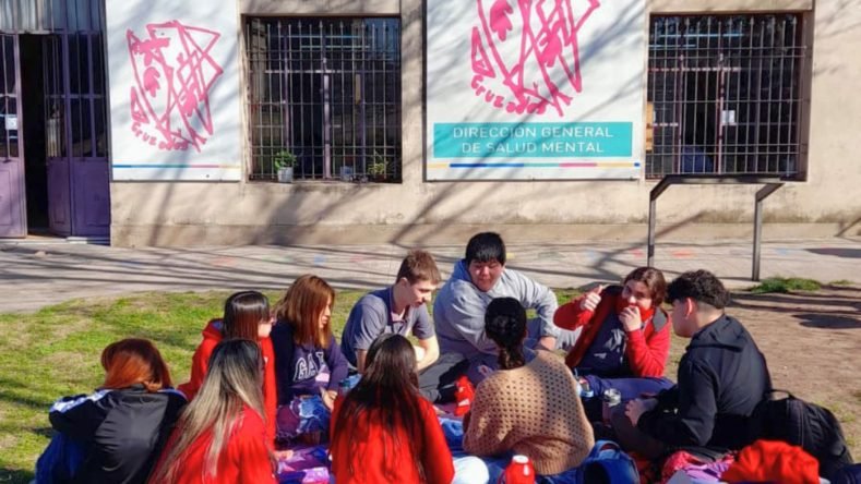 Esta imagen muestra a un grupo de jóvenes sentados en el césped frente a un edificio institucional.