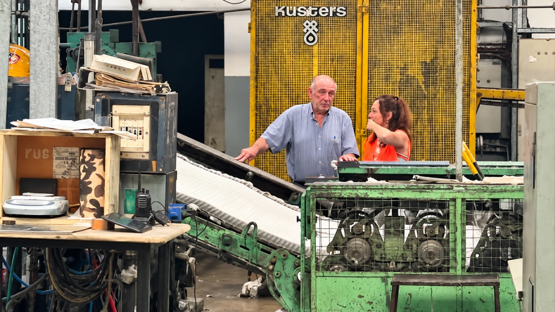 La imagen muestra el interior de una planta de producción industrial con maquinaria pesada, incluyendo un equipo que lleva la marca Küsters, visible al fondo junto a dos operarios.