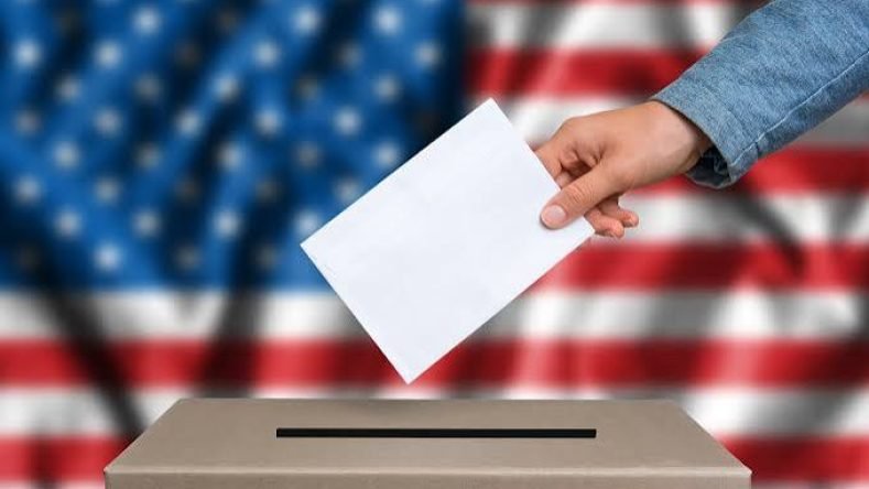 La imagen muestra una mano depositando una papeleta de votación en una urna, con la bandera de los Estados Unidos de fondo, un símbolo clásico del proceso electoral estadounidense.