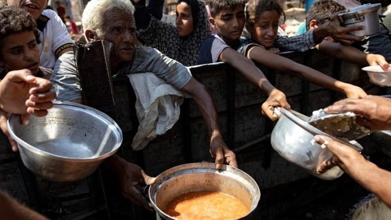 La imagen muestra a un grupo de personas, incluidos niños y adultos mayores, extendiendo ollas y recipientes vacíos para recibir comida caliente en un punto de distribución de ayuda humanitaria.