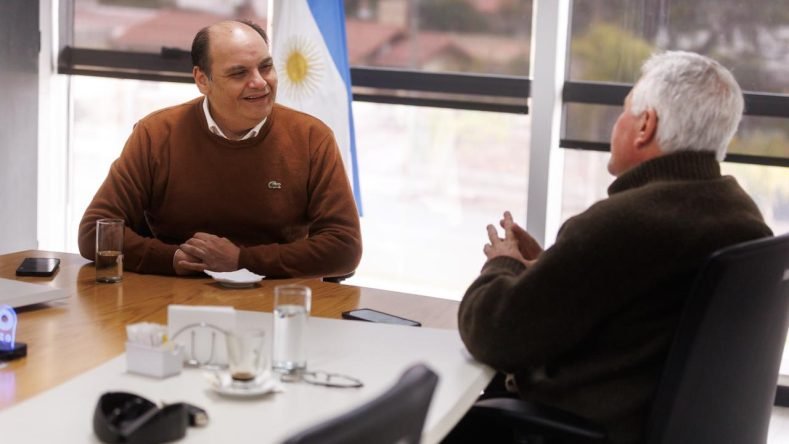 Esta imagen muestra a dos personas reunidas en una oficina frente a una mesa con vasos de agua y elementos de vajilla, con una bandera de Argentina de fondo.