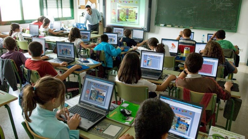 Esta imagen muestra un aula escolar moderna donde varios estudiantes están utilizando computadoras portátiles en sus pupitres frente a un profesor y una pizarra digital interactiva.