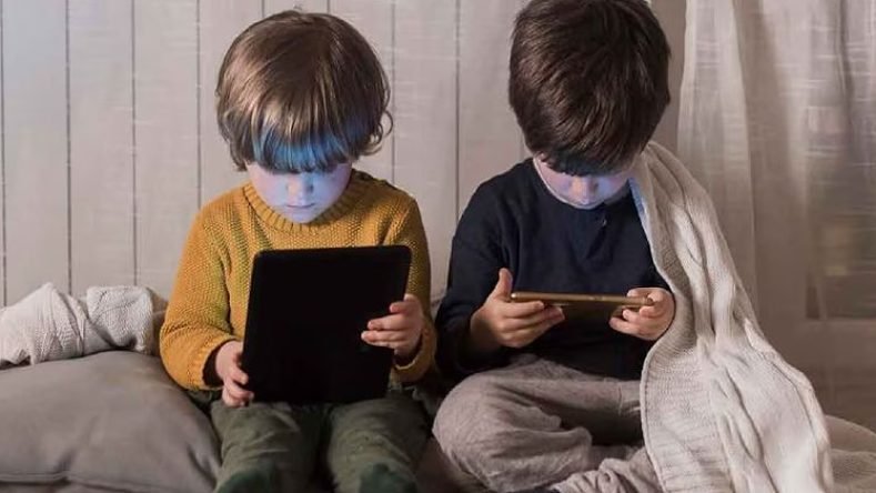 La imagen muestra a dos niños pequeños sentados en el suelo, abrigados con una manta y cojines, utilizando dispositivos electrónicos (una tableta y un teléfono móvil) con la luz de las pantallas iluminando sus rostros.