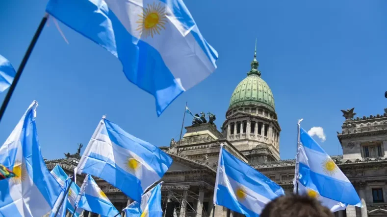 La imagen muestra el icónico Palacio del Congreso de la Nación Argentina, ubicado en la ciudad de Buenos Aires, con varias banderas argentinas ondeando en primer plano durante una manifestación o acto público.