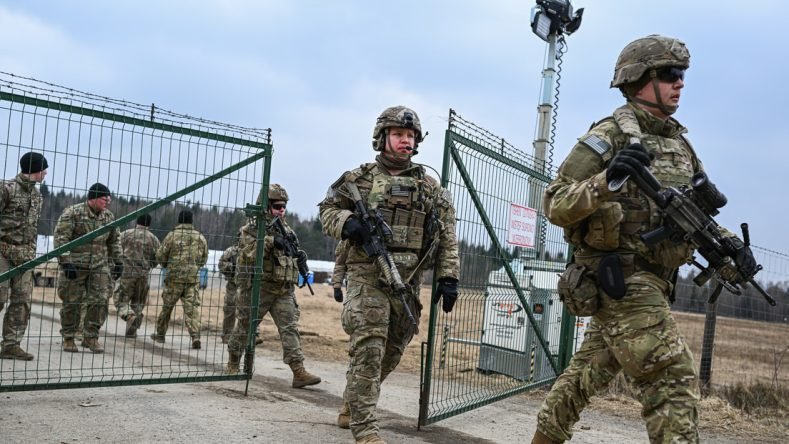 La imagen muestra a soldados del Ejército de los Estados Unidos con equipo de combate completo, incluyendo cascos, chalecos tácticos y rifles de asalto, pasando por una puerta metálica en un entorno exterior.