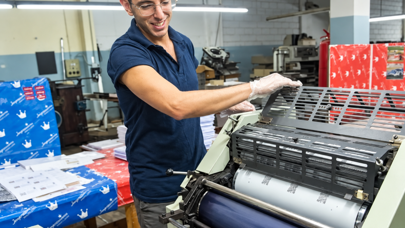 La imagen muestra a un trabajador operando una máquina de impresión offset en el marco de la labor productiva de una cooperativa o empresa recuperada.