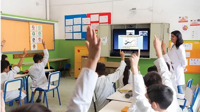 Esta imagen muestra un aula escolar con varios estudiantes sentados en sus pupitres con el brazo en alto y una docente de pie junto a una pantalla interactiva.