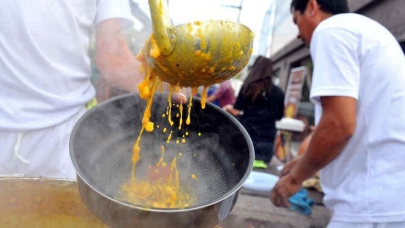 Se observa a una persona sirviendo locro, un guiso tradicional argentino, con un gran cucharón de metal desde una olla humeante.