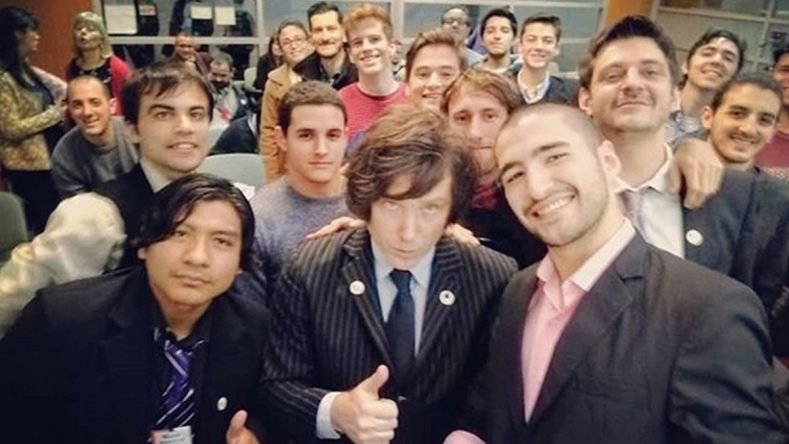 Fotografía en plano medio de un grupo de jóvenes sonrientes rodeando a Javier Milei en un aula o salón de reuniones. En el centro, Milei viste un traje oscuro a rayas, camisa clara, corbata oscura y mira fijamente a la cámara mientras hace el gesto de aprobación con el pulgar levantado. A su lado, varios jóvenes con trajes formales y vestimenta casual sonríen para la selfi colectiva. El ambiente refleja un encuentro de militancia política juvenil.