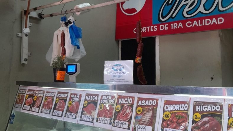 La imagen muestra el mostrador de una carnicería de barrio en Argentina con carteles de precios para diferentes cortes de carne y embutidos.