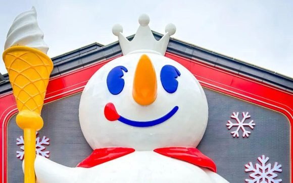 La imagen muestra la fachada de la tienda insignia de Mixue Bingcheng (蜜雪冰城) en Nankín (Nanjing), China, la cual destaca por una enorme escultura de dos pisos de altura de su mascota, el Rey de las Nieves (Snow King), sosteniendo un cono de helado.