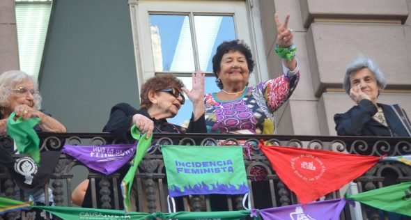 La imagen muestra a referentes históricas del movimiento feminista y de derechos humanos —como Nelly Minyervsky, Dora Barrancos y Marta Alanís— asomadas a un balcón sobre la Avenida de Mayo durante la movilización por el Día Internacional de la Mujer (#8M).