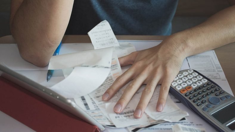 La imagen muestra a una persona estresada calculando cuentas y organizando numerosos tickets de gastos sobre un escritorio junto a una calculadora científica y una tablet.