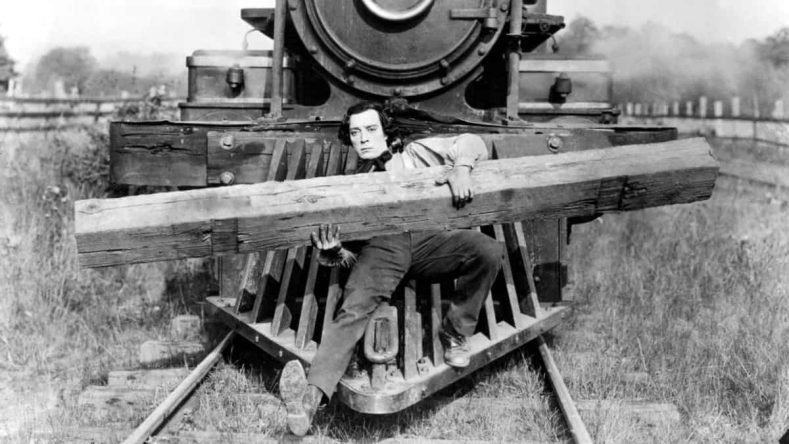 La imagen muestra al actor Buster Keaton sentado sobre elaparte delantera de una locomotora sosteniendo un pesado madero en la clásica comedia de cine mudo El maquinista de La General (1926).