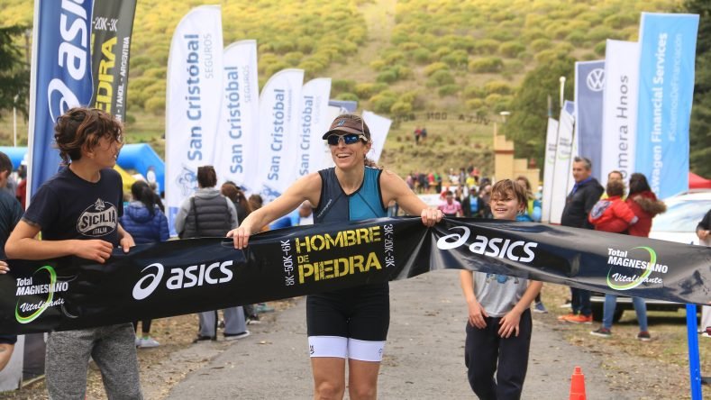 Esta imagen muestra a una participante sonriente cruzando la meta con una cinta de llegada de la competencia de duatlón Asics Hombre de Piedra en Tandil, Argentina.
