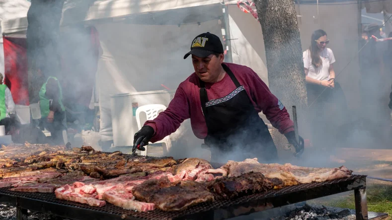 La imagen muestra a un asador con gorra negra y remera de manga larga roja manipulando carne sobre una parrilla metálica al aire libre, rodeado de humo.