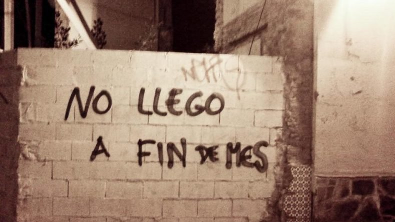 La imagen muestra una pintada callejera en letras negras sobre un muro de bloques de cemento que dice la frase popular "No llego a fin de mes".