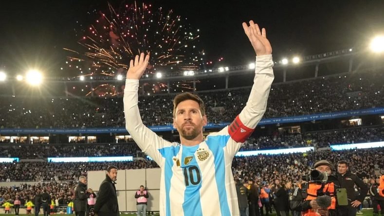 Esta imagen muestra a Lionel Messi saludando emocionado al público en el Estadio Monumental de Buenos Aires tras un partido de la Selección Argentina, con fuegos artificiales iluminando el cielo nocturno de fondo.