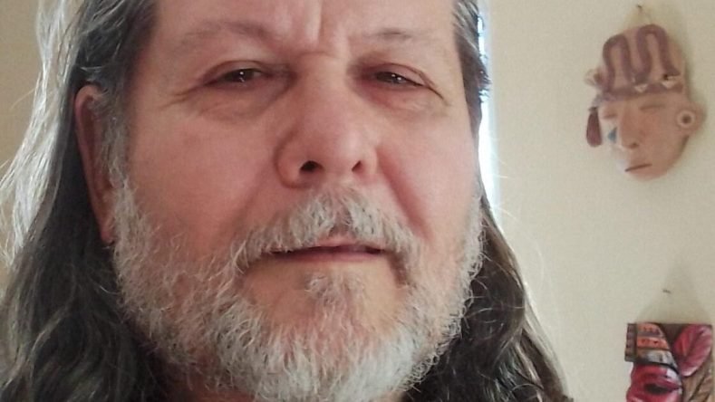 Esta imagen muestra a una persona con cabello largo y canoso y barba, vistiendo una camiseta negra frente a unas máscaras decorativas en la pared.