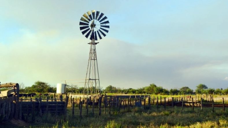 La imagen muestra un paisaje rural típico de la llanura argentina, caracterizado por un molino de viento metálico de multipalas usado para la extracción de agua subterránea.