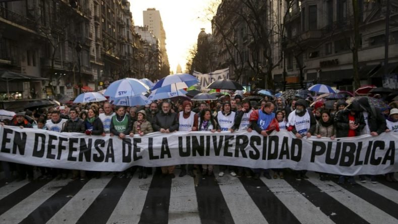 La imagen muestra una masiva movilización callejera en Buenos Aires, Argentina, encabezada por una bandera con la leyenda "En defensa de la universidad pública", en el marco de los reclamos presupuestarios y salariales del sector educativo.