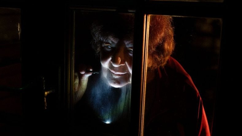 Esta imagen parece mostrar a una persona mayor asomándose a través de una ventana oscura mientras sostiene una pequeña luz, un momento que se asemeja a una escena dramática o teatral.