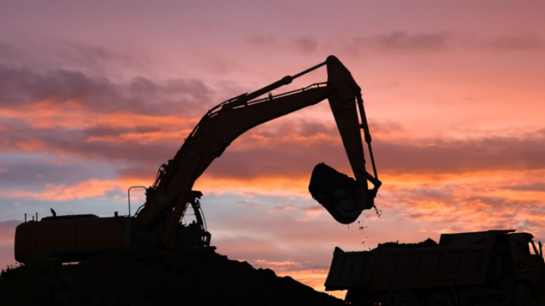 La imagen muestra la silueta de una excavadora pesada cargando tierra o arena en un camión volquete durante el amanecer o el atardecer.